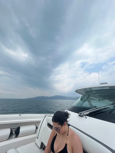 The image shows a woman seated on the deck of a white boat with a grey stripe. She is wearing a black bikini top with gold rings on the straps. Her hair is pulled back and she is wearing glasses. The boat is sailing on the blue water and there are mountains in the distance. The sky is partly cloudy with a few patches of blue sky visible. The location is Playa Herradura, Costa Rica, based on the coordinates provided. The woman appears to be looking down at something on the boat.
