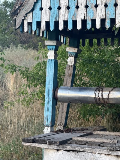 The image shows a close-up view of an old, weathered wooden well with peeling blue and white paint, located in the village of Shulhynka, Ukraine. The well structure is ornate, featuring decorative wooden elements with pointed edges. A large, metallic pipe, likely part of the well mechanism, extends horizontally across the frame, partially wrapped with a rusty chain. The pipe is supported by wooden beams. The well is situated outdoors amidst dry, tall grass and green foliage, suggesting a rural or overgrown setting. The overall impression is one of age and disuse, with the natural environment encroaching on the man-made structure.