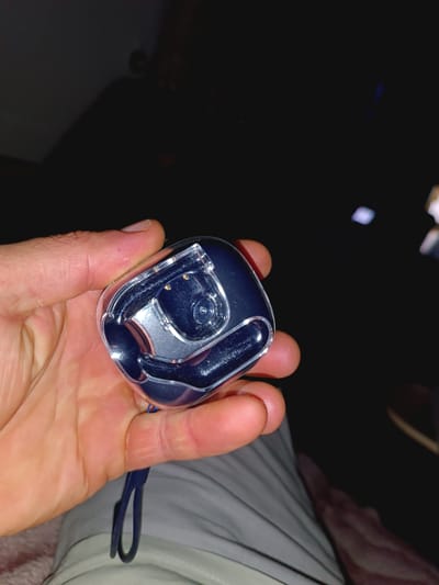 A hand in Talas City, Kyrgyzstan, holds a transparent, dark blue earbud case. The case is partially open, revealing the dark blue earbuds nestled inside. A thin, dark blue lanyard is attached to the case and drapes downwards. The background is dark and out of focus, with faint reflections of light that suggest an indoor environment with artificial lighting. The hand's skin appears dry with visible fingerprints and subtle abrasions.