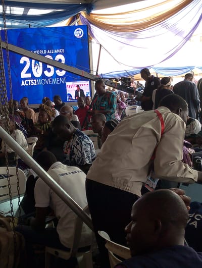 The image captures an indoor event taking place under a large tent or marquee in Dushepe, Nigeria, likely during daylight hours. The ceiling is adorned with draped fabrics in shades of blue, white, and brown/gold, supported by visible metal beams and chains. A large audience of adults is seated in rows of white plastic chairs, with many individuals visible from the back or side.
At the front of the gathering, a prominent blue LED screen displays the text "WORLD ALLIANCE," "2030," and "ACTS2MOVEMENT." A smaller inset screen on the main display shows a male speaker delivering a presentation. Among the attendees, a person is operating what appears to be a video camera on a tripod, indicating media coverage of the event. In the foreground, a man wearing a beige jacket and a red lanyard with a "MEDIA" badge is moving through the space.
Attendees are dressed in a variety of attire, ranging from casual wear to patterned traditional garments. On the back of a white shirt worn by a seated man, the text "CELEBRATING THE THREING OK" is partially legible. The visible text on the screen, along with the organized seating and media presence, suggests a formal conference, meeting, or public gathering centered around the "World Alliance 2030 Acts2Movement" initiative. The atmosphere appears attentive.