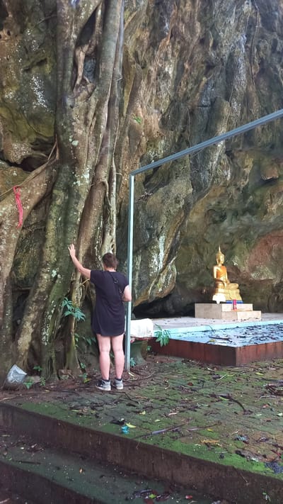 A person is standing near a cave in Ban Pak Ra, Thailand. They are wearing a dark-colored dress or shirt and sneakers. The person has one hand extended toward a large tree root system. In the background, a golden reclining Buddha statue is visible inside the cave. The cave walls are rocky and textured. The ground is covered in moss and foliage. A metal railing is also visible.
