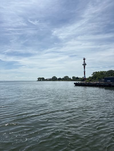A vertical shot captures a wide expanse of dark, rippling water in the foreground, with subtle white foam and reflections dotting its surface. In the mid-ground, a concrete pier juts out into the water, adorned with large, black, cylindrical fenders. Atop this pier stands a tall, red and white striped lighthouse, topped with antennas and observation decks. Behind the lighthouse, a dense line of green trees borders the land, with a glimpse of a dark-colored industrial building visible to the right of the pier. The horizon line is low, dominated by a vast sky filled with wispy, pale clouds against a muted blue backdrop. The distant shoreline is softly illuminated, with a few isolated trees and a hint of structures on a slightly elevated landmass. The overall scene depicts a serene coastal or port environment under an overcast sky.