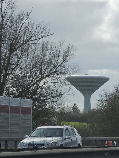 The image depicts an outdoor scene in Foetz, Luxembourg, under an overcast sky. In the foreground, a dark, blurred horizontal element occupies the bottom, above which is a grey concrete barrier with dark vertical supports. Moving into the midground, a silver Mercedes-Benz C-Class Estate (W203 generation) travels leftward, partially obscured by the foreground barrier. A red object is affixed to the vehicle's roof, and a driver is partially visible inside. To the left of the road, a grey sound barrier with a reddish-brown horizontal stripe is present. Behind the road and sound barrier, a strip of green grass is visible, bordered by numerous bare deciduous trees and some evergreen bushes. The background is dominated by the prominent, light grey water tower of Foetz, characterized by its tall, slender central column supporting a wide, disc-shaped reservoir with a ribbed underside. Additional bare trees and evergreen foliage frame the water tower. The sky is uniformly overcast with light grey clouds. No other individuals or significant actions are observable.