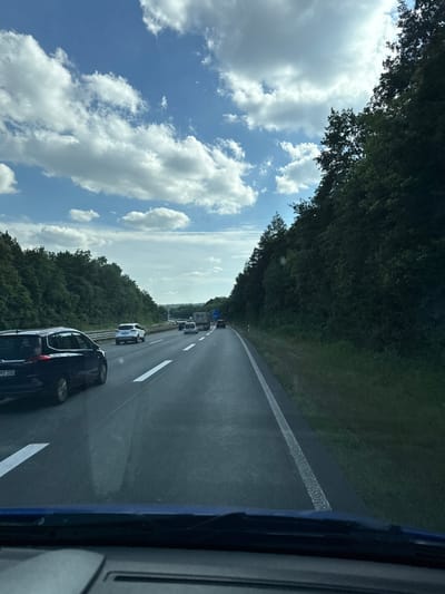 The image captures a view from a vehicle traveling on a highway in Giessen, Germany. The scene is dominated by a wide asphalt road with white lane markings, suggesting a multi-lane highway. Dense, green trees line the left side of the road, forming a thick forest. The sky above is a bright blue, dotted with fluffy white clouds, indicating fair weather.

Several vehicles are visible on the highway, indicating moderate traffic. In the foreground, the rear of a dark-colored car is prominent, with its license plate clearly visible. Further down the road, other cars and trucks are driving in the same direction. The perspective suggests the photo was taken during daylight, likely midday or afternoon, given the bright illumination and clear sky.

The overall mood is one of travel and everyday life on a German autobahn. There are no people clearly discernible in the image, as the focus is on the road and vehicles. The setting is clearly a highway environment, with the natural landscape of trees contrasting with the man-made structure of the road. The only visible text is on the license plate of the car in the foreground, which reads "MS 100".