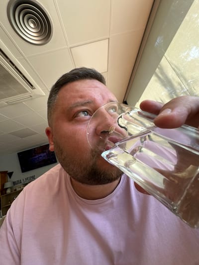 A man in a light pink shirt is drinking from a clear glass. The perspective is from below, looking up at him. The background includes a white tiled wall, an air vent, and a window with natural light. A television and some shelving are visible behind him. The location is Gurb, Spain.