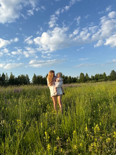 A woman is holding a toddler in a field of tall green grass with yellow and purple wildflowers. The woman has long blonde hair and is wearing a white t-shirt and shorts. The toddler is wearing a white and grey patterned dress. In the background, there is a line of trees and a blue sky with white clouds. The scene is set in the city of Koloksha, Russia. The lighting suggests it is late afternoon.