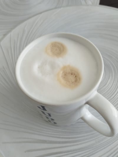 A close-up, overhead shot shows a white ceramic mug filled with frothed milk coffee. Two distinct circular patches of darker coffee foam are visible on the surface of the milk. The mug has a white handle and appears to have some dark lettering partially visible on its side, with the letters "JUG" discernable. The mug rests on a white, textured surface that resembles a placemat or a decorative plate with concentric ripple patterns. The lighting is soft, suggesting an indoor setting. There is no visible indication of the city of Novostroika, Russia, in the image.