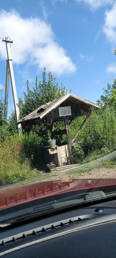 An old, weathered wooden well structure is situated amongst dense green foliage under a bright blue sky with scattered white clouds. The wellhead is covered by a gabled roof with decorative trim and appears to have sustained some damage. A metal bucket rests on the edge of the well structure, and a wooden plank leans against it. To the left of the well, a tall, concrete utility pole stands with wires attached. In the foreground, the hood and windshield wipers of a red vehicle are visible, suggesting the photo was taken from inside the car. A sign with text in Russian is affixed to the well structure. The overall scene depicts a rural or semi-rural environment. The location is the city of Puchezh, Russia.