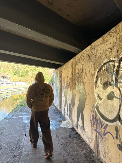 An individual is depicted from behind, walking through a concrete underpass in Wyre Forest, United Kingdom. The person is wearing a tan hooded sweatshirt with the hood up, featuring a dark, circular maze-like symbol on the back, along with loose-fitting dark brown trousers and light-colored shoes. The underpass ceiling is dark grey with visible structural beams. To the right, a concrete wall is heavily marked with peeling, discolored paint, dark water streaks, various spray-painted graffiti tags in black, purple, and white, and several stylized dark silhouette figures. A crumpled white plastic bag rests on the ground at the base of this wall. The concrete pathway is damp in areas and littered with debris. Ahead, the underpass opens to a brightly lit exterior, revealing a calm canal or waterway reflecting the sky, parallel railway tracks on the far bank, a small light-colored building, and abundant green trees under a clear sky. The individual is moving towards this open, illuminated area.