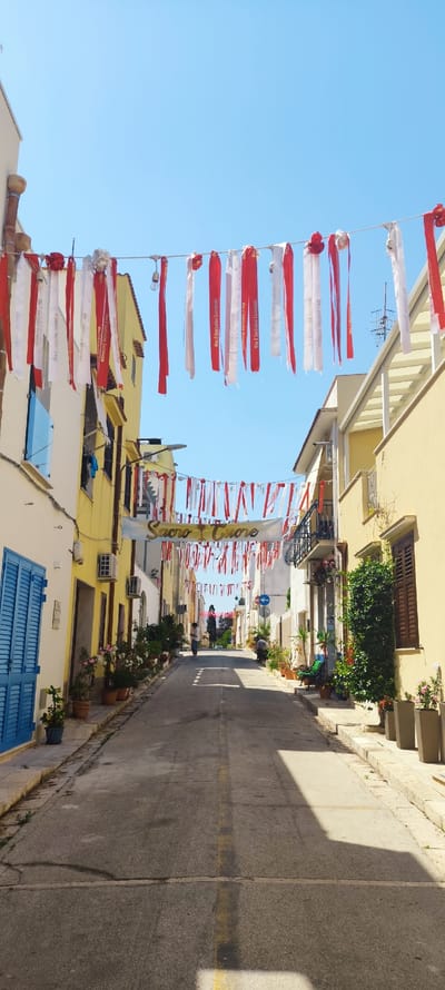 The image depicts a sun-drenched street in San Vito Lo Capo, Italy, adorned with red and white streamers. The narrow street is lined with buildings, primarily painted in yellow and white, with blue shutters on one side and brown wooden shutters on the other. Potted plants and greenery are placed along the sidewalks and near the buildings, adding a touch of natural beauty to the urban setting.
Several people are visible walking along the street. Two individuals are in the mid-ground, walking away from the camera, and another person is seen further in the distance, also walking. The activity appears to be a festive occasion, as indicated by the decorations strung across the street and a banner overhead.
The banner, with gold lettering on a white background, displays the words "SACRO CUORE" which translates to "Sacred Heart." Some of the streamers also have text on them, appearing to be repeating phrases. The bright sunlight and clear blue sky suggest it is a warm, sunny day, likely in the late morning or early afternoon. The scene evokes a sense of celebration and a peaceful, traditional Italian street atmosphere.