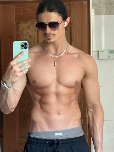 A male subject is taking a mirror selfie in a room. He is shirtless, wearing sunglasses, a pearl necklace, and grey boxer briefs with black shorts pulled down over them. He has dark hair pulled back into a bun and a mustache. He is holding a light blue smartphone in his left hand. A watch is visible on his left wrist. The background consists of a wooden door on the left and a tiled wall on the right. A digital display is visible on the tiled wall. There is no indication of the city of Borowa, Poland.