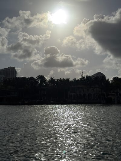 The image presents a waterfront scene, likely in Bal Harbour Village, Florida. The right side of the image is dominated by a body of water, possibly the Intracoastal Waterway, with shimmering reflections of sunlight off the surface. On the opposite bank, a silhouette of palm trees and buildings is visible, indicating the location of residential or commercial properties. The sky is overcast with clouds and the sun is shining. A construction crane is silhouetted amid the foliage and buildings. The lighting suggests either early morning or late afternoon. The buildings closest to the water’s edge are obscured in shadows. A taller building can be made out further in the background.