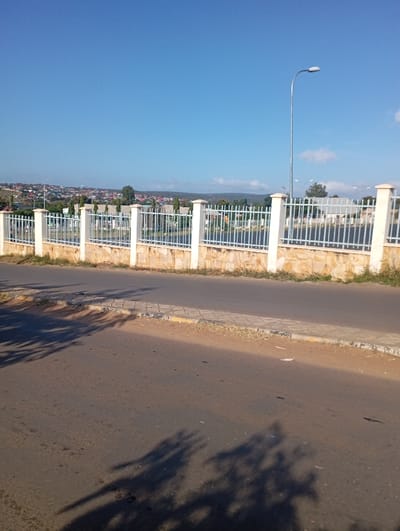 This eye-level outdoor shot depicts a sunny street scene, likely in Namanga, Kenya. The foreground features a wide, dusty asphalt road with prominent, long dark shadows stretching across it from left to right, suggesting either early morning or late afternoon. A light-colored concrete curb, partially marked with yellow, separates the road from a narrow strip of dirt and sparse grass on the right.
In the midground, a white metal picket fence with robust stone pillars runs horizontally across the frame. Behind this fence, a residential area is visible, characterized by numerous houses and buildings, many with distinctive red roofs, nestled among green trees and other vegetation. A tall, silver street lamp with a curved arm stands near the right side of the fence, reaching into the sky.
The background is dominated by a clear, bright blue sky with only a few faint, wispy clouds. A distant, gently rolling ridge or hill line defines the horizon. The scene conveys a sense of calm in a typical urban or semi-urban environment on a bright, clear day. There are no people visible, no discernible activity taking place, and no visible text in the image.