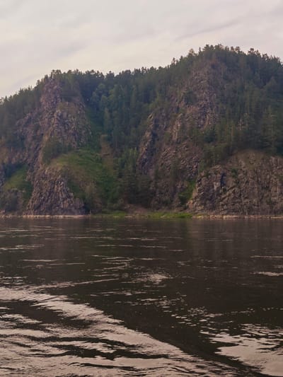 A wide, dark river occupies the foreground of the image, with ripples and waves reflecting the overcast sky. In the middle ground, a steep, rocky cliff face rises directly from the riverbank. The cliff is partially covered in dense green vegetation and scattered trees. The upper portion of the cliff is densely forested with dark green coniferous trees. The sky above is a pale gray, suggesting a cloudy day. The scene appears to be a natural landscape, likely in a rural or wilderness area, with no visible signs of human habitation or activity beyond the implied presence of a river suitable for travel. The location is identified as Beregovaya-Podemnaya, Russia.