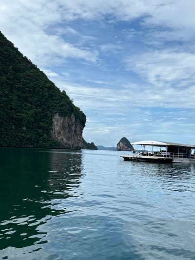 A wide shot captures a serene seascape in Khlong Khian, Thailand, under a cloudy sky. On the left, a colossal, verdant limestone karst rises steeply from the dark green water, its sheer, craggy face a dramatic contrast to the lush vegetation covering its slopes. The water in the foreground is calm, reflecting the sky and the imposing cliffs.

To the right, a floating platform, possibly a restaurant or a tour boat, is anchored in the lighter blue water. It has a white canopy covering its roof and appears to be equipped with seating and tables. A few individuals are visible on the platform, including one person wearing a green shirt standing near the railing. In the distance, another smaller, cone-shaped karst formation juts out of the sea, adding to the picturesque landscape. The overall scene evokes a sense of tranquility and natural beauty.