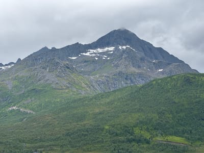 The image shows a mountain landscape under an overcast sky. The foreground is dominated by a densely wooded, green hillside. A winding path or road is visible on the left side of the wooded slope, and further to the right, there is a power line with a pylon. The middle ground features a rocky, grey mountain with patches of snow near its peak. The mountain has steep, rugged slopes. The background consists of a cloudy sky. The location is identified as Langvassbukta, Norway. There are no people or animals visible in the image.