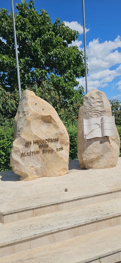 The image depicts a monument in Cserszegtomaj, Hungary, dedicated to an event in 1956. The monument consists of two large, light-colored rocks. The rock on the left has text engraved on it, reading "IN MEMORIAM 1956 ALLITVA 2006-BAN". The rock on the right features a stylized carving of an open book with a curved spine, adorned with a ribbon. The monument is situated on a stone staircase. Behind the monument, there is lush green foliage and a clear blue sky with scattered clouds. A small rodent is visible on the stone steps in the foreground.