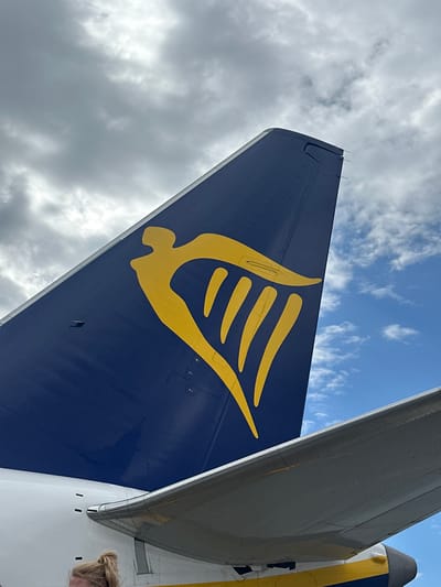 The image captures a close-up view of the tail fin of a Ryanair airplane, prominently displaying the airline's yellow harp logo against a dark blue background. The tail fin is positioned against a sky filled with a mix of white, fluffy clouds and darker, overcast clouds. Below the tail fin, a portion of the airplane's wing is visible, appearing metallic gray and reflecting some of the sky's light. A section of the airplane's fuselage, painted white with yellow and blue stripes, is also visible at the bottom of the frame. A woman with blonde hair tied up in a bun is partially visible at the very bottom of the image, suggesting the photo was taken from ground level. The scene is consistent with an airport environment, and given the detail, it is likely taken at John Paul II International Airport Kraków–Balice (KRK) in Balice, Poland.