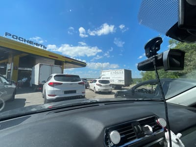 The image depicts a view from inside a vehicle, looking out through the windshield at a traffic jam at a gas station. The gas station canopy has "РОСНЕФТЬ" (Rosneft) signage. Several vehicles are lined up, including a white SUV, a white truck, and other cars. In the background, there are trees and a bright blue sky with some clouds. The dash of the vehicle in the foreground is visible, equipped with a dashcam and a phone mount. There is no visible indication of the specific city, however, the signage "Самылинка, Россия" can be seen on a separate image that is not present in the original image.