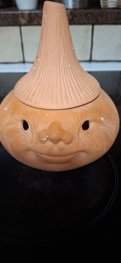 The image shows a terracotta mushroom-shaped object with a face sculpted onto its cap. The object has a wide, round cap with two circular eye holes and a sculpted nose and mouth, giving it a whimsical expression. The stem of the mushroom is ridged, resembling the gills of a mushroom or perhaps the texture of carved wood. The terracotta is a warm, earthy orange color and has small white speckles scattered across its surface. The object appears to be resting on a black stovetop, which has subtle circular markings indicating burner elements. In the background, out of focus, are tiled surfaces and what looks like kitchen cabinetry. The lighting suggests an indoor setting, likely a kitchen, with no specific indication of the time of day or weather. The object itself is the sole focus, and no people are visible. There is no visible text in the image. The overall impression is of a decorative garden ornament or a playful piece of kitchenware, possibly intended for storing something or as a quirky planter. The location context provided, Orimattila, Finland, doesn't offer any visual cues within the image itself but indicates the geographical setting where the photo was taken.