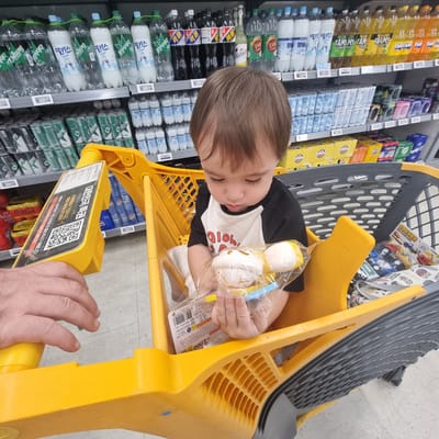 The image shows a young child, likely a toddler, sitting in a yellow shopping cart in a grocery store aisle. The child is wearing a black and white raglan t-shirt with a graphic design. The child is holding a toy that is partially visible inside a clear plastic bag. The shopping cart is filled with various items in the background, including bottled drinks on shelves, cans, and boxes of goods. A person's hand is visible on the left side of the image, gripping the handle of the shopping cart. The environment appears to be a well-lit supermarket. Based on the product packaging and price tags in Korean, this scene is taking place in Wonju-si, South Korea.