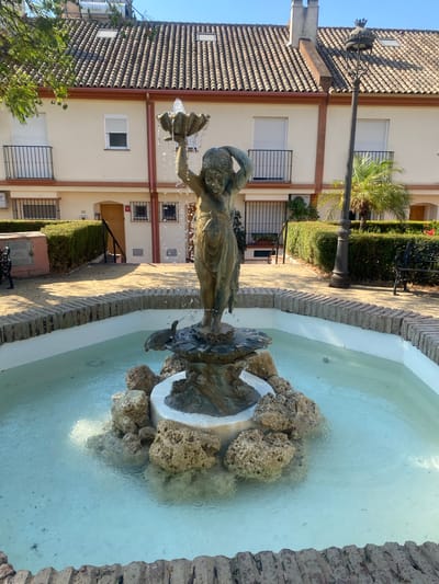 This is a photograph of a bronze statue of a young woman holding a shell above her head, with water spraying from the shell. The statue stands in the center of a circular fountain filled with clear blue water. The fountain is surrounded by a low brick wall and decorative rocks. In the background, there are buildings with terracotta tile roofs and white facades, typical of Spanish architecture in the town of Cancelada, Spain. The buildings have balconies, windows, and doors. A lamppost and some manicured hedges are also visible. The overall scene depicts a tranquil public space.