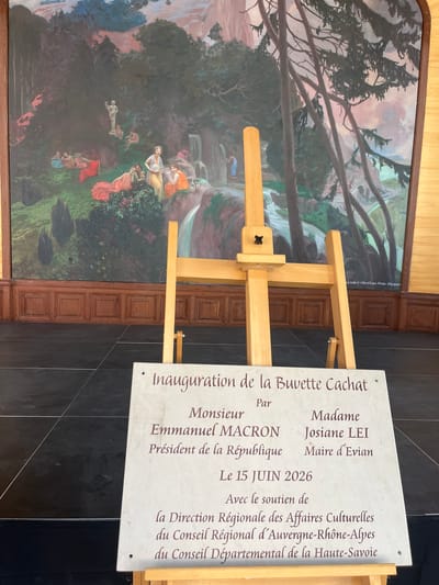 The image shows a plaque on an easel in front of a mural in Évian-les-Bains, France. The plaque reads "Inauguration de la Buvette Cachat" (Inauguration of the Cachat Drinking Fountain) and lists attendees and sponsors. The mural depicts a landscape with figures in classical attire, possibly a scene from mythology or historical times. The easel is made of wood, and the plaque is stone with engraved text. The background includes a wooden paneled wall and a tiled floor. The setting appears to be an indoor or covered outdoor space, likely related to a public event.
