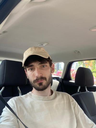 The image shows a young man wearing a beige t-shirt and a beige baseball cap, sitting in the passenger seat of a car. He has brown hair and eyes, and a beard. The car's interior is visible, including the headrests and the ceiling. Outside the window, buildings and trees can be seen, suggesting an urban environment. The lighting indicates it is daytime. The street sign visible outside the window is in Polish and reads "STOLOKOWSKIEGO". Based on this, the location is Stolokowskego Street in Bielsko-Biala, Poland.