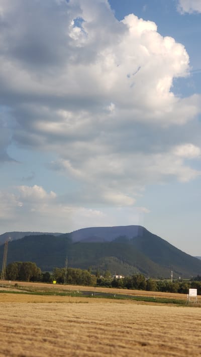 The image shows a rural landscape with a field of dry, golden grass in the foreground. In the middle ground, there is a line of trees and a road. In the background, there are rolling hills covered in dark green forests. The sky is partly cloudy with large white clouds against a blue sky. There are no people or animals visible in the image. The scene is peaceful and appears to be taken during daylight. There is no specific information about the location in the city of Beňadiková, Slovakia, within the image itself.