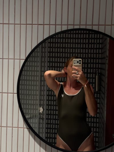 A woman in a dark green one-piece swimsuit with white trim is taking a mirror selfie in what appears to be a bathroom. She is standing in front of a circular mirror that reflects a patterned tiled wall behind her. The tiles on the wall in front of her are elongated and white with thin pink grout lines. She is holding her phone up to take the picture with her right hand, and her left hand is adjusting her hair. She is wearing a ring on her right hand and a bracelet on her left wrist. A silver soap dispenser is visible in the reflection of the mirror. The image does not contain any contextual information that would indicate the location is in Kaji-Say, Kyrgyzstan.