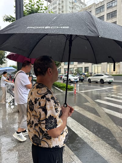 In Xuhui District, China, on a rainy day, several individuals are gathered near a crosswalk under umbrellas. A man in the foreground, facing to the right, holds a black umbrella with the "PORSCHE" logo visible and a cigarette in his right hand. He is wearing a cream-colored shirt with a black floral pattern and black pants. Behind him, another man in a white t-shirt and glasses is holding a dark umbrella. Further back, a woman in a white top and light-colored pants is pushing a shopping cart, holding a light blue umbrella. Cars are driving on the wet street, and buildings are visible in the background. The crosswalk markings are clearly visible, with raindrops creating ripples on the wet pavement.