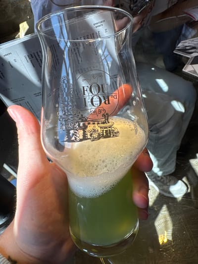 A person holds a glass of green liquid with white foam, obscuring a partial view of a table menu. The menu lists spirits and aperitifs, including absinthe, gin, vodka, and Ricard. The glass has the logo "Du R Fou" printed on it. Partial view of another person's legs and a second menu are visible. The setting appears to be outdoors in bright sunlight. There's no explicit information to confirm the location in Epagny, Switzerland.