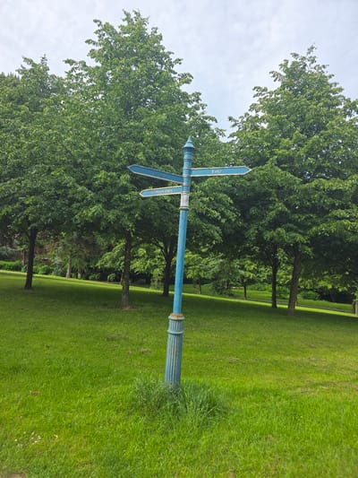 A blue directional signpost stands in a grassy park area in Stockton-on-Tees, United Kingdom. The signpost has three teal-colored directional arrows. The top arrow points left and reads "Park Fold". The middle arrow points right and reads "Lake". The bottom arrow points left and reads "Barden Gates". The signpost is weathered and has some rust. It is positioned in front of a dense line of green trees. The ground is covered in a lush green lawn with some patches of darker grass. The sky is overcast with some white clouds. The overall scene depicts a peaceful park setting.