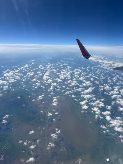 The image is taken from an airplane window, looking down at a vast landscape below. The perspective is tilted, with the airplane wing prominently visible in the foreground on the left side of the frame. The wing is white with a red and black tip, suggesting it's part of a commercial aircraft.

Below the wing, the scene is divided into distinct layers. The upper left is dominated by a deep, clear blue sky, transitioning into a hazy, lighter blue near the horizon. A thin band of white clouds stretches across the horizon.

The majority of the image is a view of the earth from high altitude, dotted with numerous fluffy white cumulus clouds casting shadows on the land below. The landscape beneath the clouds is a patchwork of green fields, possibly farmland, interspersed with numerous small bodies of water, likely lakes or ponds. A prominent, winding river with darker water flows through the scene, with tributaries branching off. There are signs of human habitation, with small clusters of buildings visible, suggesting villages or towns. Roads and pathways crisscross the terrain. The overall impression is one of a rural, possibly somewhat remote, location.

The lighting suggests it is daytime, with the sun high enough to cast clear shadows from the clouds. The weather appears fair, with scattered clouds and clear patches. There is no visible text in the image. Given the location context provided as Ragozina, Russia, this depicts a view over that general region. The activity is passive observation from an aircraft in flight.