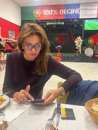 A woman with medium-brown hair, wearing rectangular glasses and a dark purple sweater, is seated at a table in a Portuguese cultural establishment in Décines-Charpieu, France. She is focused on a black smartphone held horizontally in both hands. Her visible right hand displays light pink fingernails and wears two rings. A metallic wristwatch is visible on her left wrist. On the white-tableclothed table in front of her, partially visible elements include a plate with food (containing fries), a silver fork, a black-handled knife, and a black wallet displaying a red Mastercard. A woven bread basket is positioned to the right. The background features a stage area adorned with Portuguese flags and banners. A large banner prominently reads "100% DÉCINOIS" alongside a Portuguese flag. Below this, the word "DÉCINES" is displayed within a heart shape encompassing the Portuguese flag. To the right of the stage, a large screen exhibits a soccer match. The environment is decorated with artificial flowering branches and hanging faux greenery.