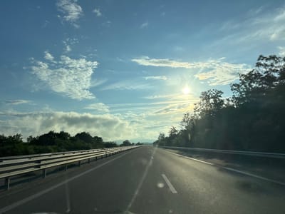 The image depicts a road, likely a highway, stretching towards a distant horizon under a partly cloudy sky. The sun is visible and shining brightly, casting a warm glow and creating lens flares on the road surface. On either side of the road, there are guardrails and dense green trees. The road itself is asphalt with clear white lane markings. The sky features a mix of scattered white clouds and wispy cirrus clouds against a pale blue backdrop. The scene appears to be captured during the daytime, possibly late afternoon or early morning due to the angle of the sun. The location of Yazdach, Bulgaria is not identifiable from the visual information provided in the image.
