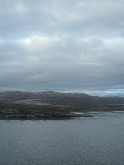 The image displays a panoramic view of a rugged coastal landscape under an overcast sky, located in the vicinity of Stanley, Falkland Islands (Islas Malvinas). The upper half of the frame is dominated by a uniform, dense grey cloud cover. Below this, a calm body of dark grey-blue water occupies the foreground, exhibiting subtle ripples across its surface. The water meets an irregular, rocky shoreline that stretches horizontally across the mid-ground. Immediately behind the shoreline, the land appears sparsely vegetated with dark, low-lying scrub or peat. Further inland, a series of low, undulating hills are visible, characterized by prominent light-grey rock formations interspersed with patches of dark vegetation. No individuals, man-made structures, or active elements are present in the scene.