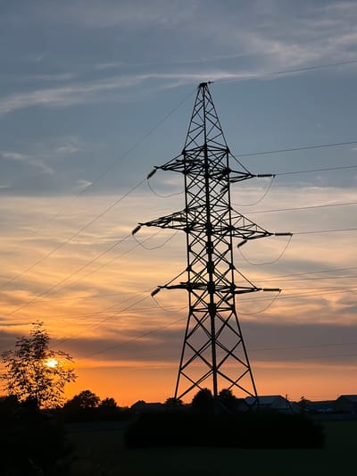 The image depicts a silhouetted power tower against a sunset sky in the village of Yasenivtsi, Ukraine. The sky is a gradient of orange and blue, with thin clouds streaking across. Several power lines stretch horizontally across the frame, supported by the tower. In the foreground, the silhouette of a tree with the sun peeking through its leaves is visible on the left. The bottom of the image shows a dark silhouette of the landscape, including trees and some buildings. The scene is peaceful and atmospheric, capturing the natural beauty of a sunset juxtaposed with industrial infrastructure.