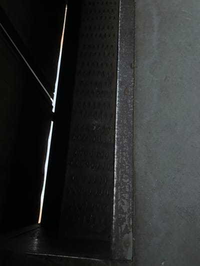 The image shows a close-up view of a dark, metallic structure with a textured surface on the left, juxtaposed with a smoother, gray metallic surface on the right. A bright, vertical sliver of light pierces the darkness on the far left, suggesting an opening or gap. The textured surface on the left appears to be made of metal with a grid of small, raised, rectangular bumps or patterns, and is slightly worn and aged. A darker, slightly rusty metallic strip runs vertically along the edge between the textured and smooth surfaces. The smooth gray surface on the right is somewhat grainy and appears to be a flat panel or wall. The overall scene suggests an industrial or utilitarian environment. Without further context, it is impossible to definitively place this image in Hlevakha, Ukraine, or provide details about people, actions, or interactions.