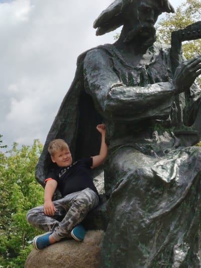 A young boy is seated on a stone next to a large bronze statue of a man playing a musical instrument. The boy is wearing a black t-shirt with orange trim, camouflage pants, and blue shoes. He is looking towards the camera and has his left arm raised. The statue is of a man with a beard and a hat, wearing a long robe. He is holding a stringed instrument. The background consists of green trees and a cloudy sky. The location is the monument to Pyotr Tchaikovsky in the village of Balyko-Shchuchynka, Kyiv Oblast, Ukraine.
