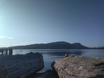 This image depicts a serene waterscape with mountainous terrain in the background, under a clear blue sky. The foreground is dominated by two large, rough-textured rocks. To the left, a pier extends into the calm water. In the distance, a range of hills or mountains forms a dark silhouette against the sky. A yellow buoy floats in the water between the pier and the rocky foreground. The scene is illuminated by natural daylight, suggesting it was taken outdoors in a coastal or fjord region. The overall impression is one of natural beauty and tranquility. The location is identified as Bremsnes, Norway.