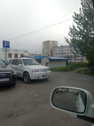 In Magadan, Russia, a white Suzuki Grand Vitara is parked on a gravel lot next to a black Audi SUV. The Suzuki has a license plate that reads "T 853 EK 49 RUS." A parking sign with a wheelchair symbol is visible in the background, indicating disabled parking. Behind the cars, there are several buildings, including a multi-story apartment complex and a smaller, shed-like structure with a blue roof. The sky is overcast, suggesting a cloudy or rainy day. The overall scene appears to be a residential or commercial area in the city.