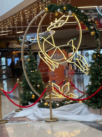 This image depicts the interior of a commercial establishment, likely a shopping mall, located in Hérouville-Saint-Clair, France, during the winter holiday season.
In the mid-ground, a large, illuminated Christmas decoration is central, featuring the outline of a reindeer within a circular frame, composed of warm white LED lights. This decoration is further adorned with green evergreen garland and spherical ornaments in gold and silver. It is flanked by two artificial evergreen Christmas trees, each decorated with lights and small ornaments. The base of this display is covered in white fabric, simulating snow, and includes a smaller, star-shaped illuminated decoration on the ground. The entire display area is cordoned off by red velvet ropes suspended between golden stanchions.
In the background to the left, a portion of a McDonald's restaurant entrance is visible, identified by a partially obscured red sign with white lettering and additional festive lighting. Two blurred human figures are discernible near what appears to be ordering kiosks or a service counter within the McDonald's area.
To the right in the background, large, colorful promotional posters featuring animated characters (including a pig and a blue humanoid figure) are displayed. Partially legible text "Disney Belles places" is visible on one poster. Beyond these posters, reflective surfaces suggest glass doors or windows.
Overhead, multiple strands of illuminated icicle-style string lights hang from the ceiling. The floor is a polished, light-colored tiled surface reflecting the ambient and decorative lighting. No explicit actions or interactions are clearly discernible from the blurred figures.