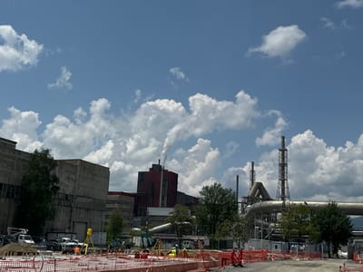 The image depicts an industrial complex in Zlatitsa, Bulgaria, under a bright, partly cloudy sky, with active construction in the foreground. In the midground and background, large industrial buildings are prominent, characterized by their concrete and reddish-brown facades. One tall, reddish structure features a chimney emitting a distinct plume of white smoke, indicating ongoing industrial operations. Numerous other tall, slender smokestacks and extensive networks of large, metallic pipes, supported by intricate scaffolding, are scattered throughout the scene.

The foreground is dominated by an active construction site. Orange safety netting and barriers define the work area, which contains piles of excavated earth and construction materials, including rebar. Several construction workers are visible, dressed in high-visibility orange and yellow vests and hard hats. One worker is operating surveying equipment mounted on a yellow tripod, suggesting precise measurement or mapping is underway. Various vehicles, including white vans and light trucks, are parked within or near the construction zone.

Interspersed among the industrial structures and construction areas are scattered green trees and bushes, providing some natural contrast to the built environment. The sky above is a clear blue, dotted with numerous fluffy white cumulus clouds, indicating a bright, sunny day, likely during midday or early afternoon. The scene conveys a sense of active industry and ongoing development. No discernible text is visible on any signs or structures.