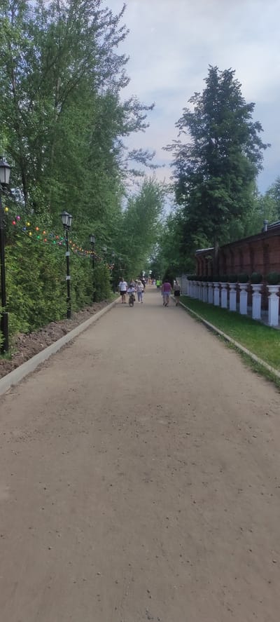 The image depicts a wide dirt path in Grebnevo, Russia, bordered by lush green trees and a low concrete curb on the left, and a decorative white balustrade and a brick wall on the right. A string of colorful lights, predominantly yellow, red, green, and blue, are strung along lampposts on the left side of the path, creating a festive atmosphere. Several people are walking and one person is on a bicycle on the path, moving away from the camera. The sky is overcast. The overall scene suggests a public park or recreational area.
