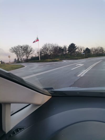 The image, captured from inside a vehicle, presents a view of a road junction or turn-off in Stefanovo, Bulgaria. A paved road stretches into the distance, featuring double solid white lines in the center and a prominent right-turn arrow painted on the right lane. On the left side of the road, a tall white flagpole displays the Bulgarian flag, characterized by its horizontal white, green, and red stripes, acting as a clear national identifier.
Bare-branched deciduous trees, indicative of late autumn, winter, or early spring, line both sides of the road and extend into the background, interspersed with some evergreen foliage. The sky above is light and overcast, suggesting either early morning, late afternoon, or simply a cloudy day, contributing to the soft, diffused light across the scene. The road surface appears damp, possibly from recent rain.
To the right of the road, partially obscured by trees, a blue directional sign with white text is visible, pointing towards "Шумен" (Shumen) and "Община" (Municipality), indicating the direction to the city of Shumen. In the foreground, parts of the car's interior, including the dashboard and windshield pillars, frame the outdoor view, confirming the perspective from within a vehicle. No specific activity beyond driving is depicted.