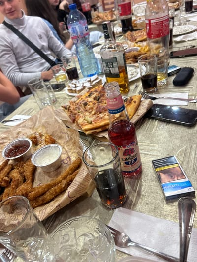 The image shows a table covered in various food and drink items, along with personal belongings, at what appears to be a social gathering.
**People:** Parts of at least two people are visible in the lower left corner. One person is wearing a white long-sleeved shirt.
**Subjects:** The primary subjects are food and drink items on the table.
**Objects:**
* Bottles: Coca-Cola, water, Somersby cider, and an unidentified brown liquid in a clear bottle.
* Food: Pizza, breaded sticks with dipping sauces.
* Glasses: Several glasses, some containing dark liquids.
* Personal items: A smartphone, a sunglasses case, and a pack of Muratti XS cigarettes.
* Cutlery: Forks and knives.
* Paper napkins.
**Actions/Interactions:** Indeterminate
**Environment:** The scene is indoors, on a table. The lighting appears to be artificial. The objects are cluttered and suggest a recent or ongoing meal.
**Location:** The presence of "0700 10 323" on the cigarette pack could suggest that the location is within Bulgaria. However, there is no direct visual confirmation of the location in Mirkovo, Bulgaria in this image.