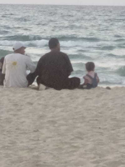 Three individuals are depicted from behind, seated on a sandy beach, facing the ocean. The scene is located in Al-Mawasi, Palestinian Territories. On the left, an adult wears a white long-sleeved shirt with a yellow daisy emblem on the back and a white baseball cap. In the center, another adult wears a dark-colored short-sleeved shirt. To the right, a small child wears light-colored overalls with visible shoulder straps. All subjects are sitting on the light-colored sand, with their backs to the viewer, gazing out at the light blue-grey ocean with small waves. The horizon is visible, indicating an open sea environment under an overcast or hazy sky.
