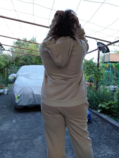 A person wearing a light tan hooded sweatshirt and matching sweatpants stands with their back to the camera, hands raised and tangled in their dark, curly hair. They are outdoors in what appears to be a yard or driveway. To the left of the person, a car is covered with a large, silver tarp, and a small grey and white cat sits on the ground in front of it. In the background, there are trees, green foliage, and some sort of metal structure. The scene is lit by natural daylight. This image was taken in Shchastia, Ukraine.