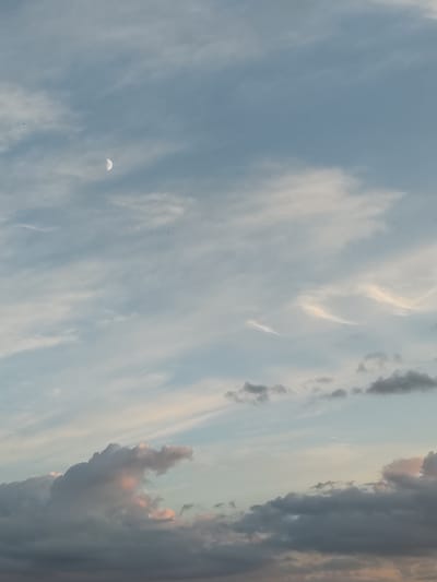 A view of the sky above the city of Chemoshur, Russia, is depicted. The sky is filled with clouds in various shades of blue, white, and gray. In the upper left quadrant of the image, a crescent moon is visible. The clouds are textured, with some appearing wispy and others more dense. There are no discernible people, subjects, objects, or actions in the frame. The scene appears to be a natural landscape photograph focused solely on atmospheric conditions.